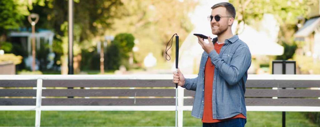 A man wearing dark glasses standing outdoors by a park bench, holding a white cane and smiling while speaking into a phone he holds in front of his mouth.