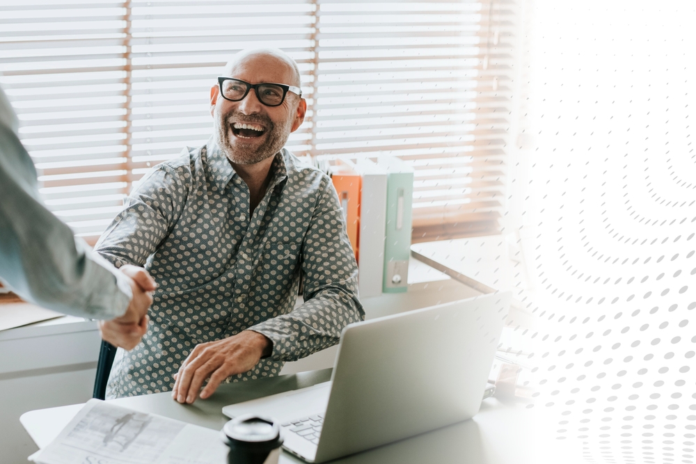 A person with a bright smile wearing thick glasses sitting at a desk with a laptop, shaking hands with someone in an office