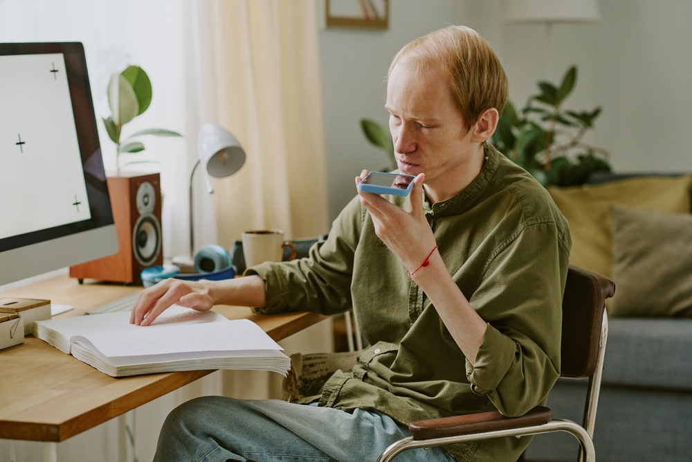 A person reading a braille book and speaking into a smartphone held close to their mouth at a desk with a computer.