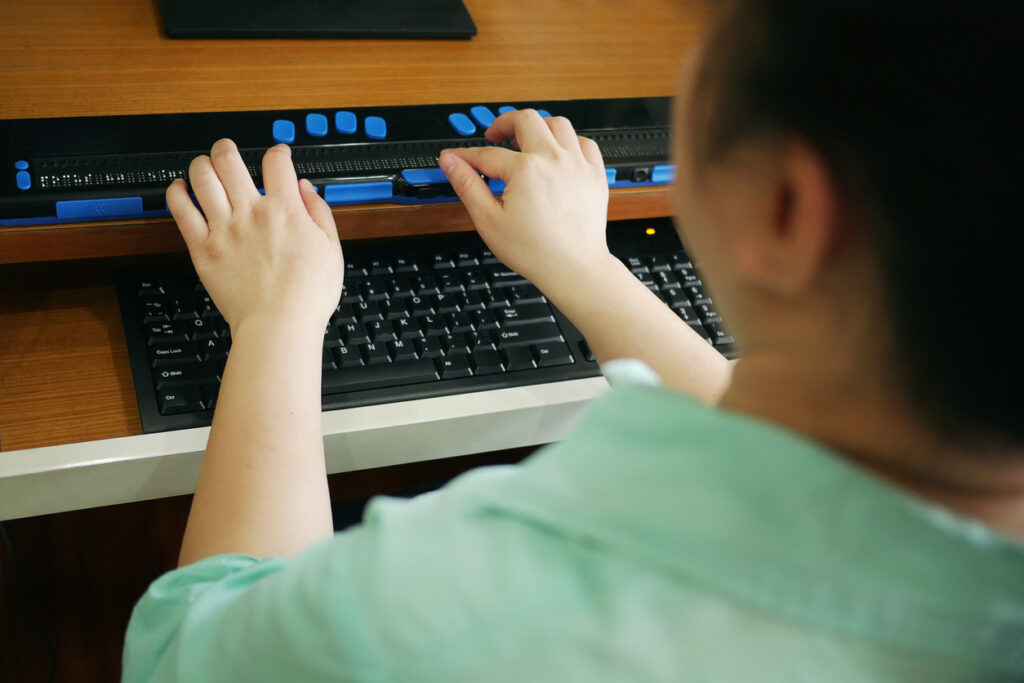 A person using a refreshable braille display alongside a computer keyboard at their workstation.