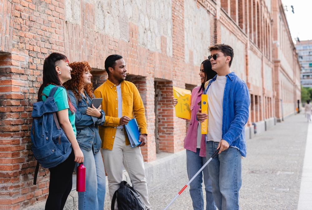 A person wearing dark glasses and holding a white cane standing outdoors talking with a group of peers beside an office building.