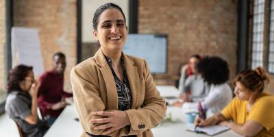 A professional with a visual disability standing and smiling confidently in a meeting room, with colleagues talking together in the background