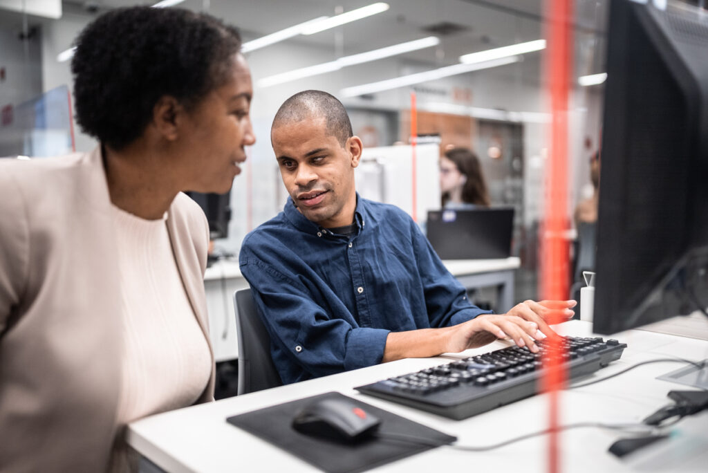 A person with a visual disability seated at a computer workstation, talking to a colleague in a modern office.