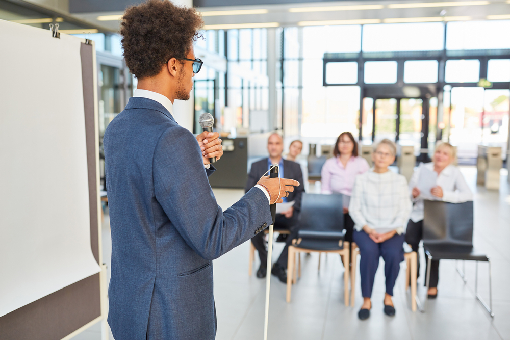 A person wearing dark glasses standing holding a white cane and speaking into a microphone in front of a seated group during a workplace training session