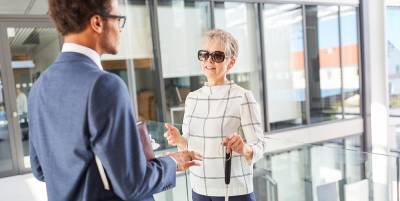 A person using a white cane while speaking with a coworker in a bright office hallway