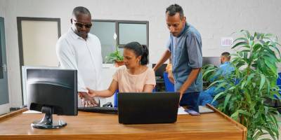 A person wearing dark glasses standing beside two coworkers as one guides their hand on a computer keyboard