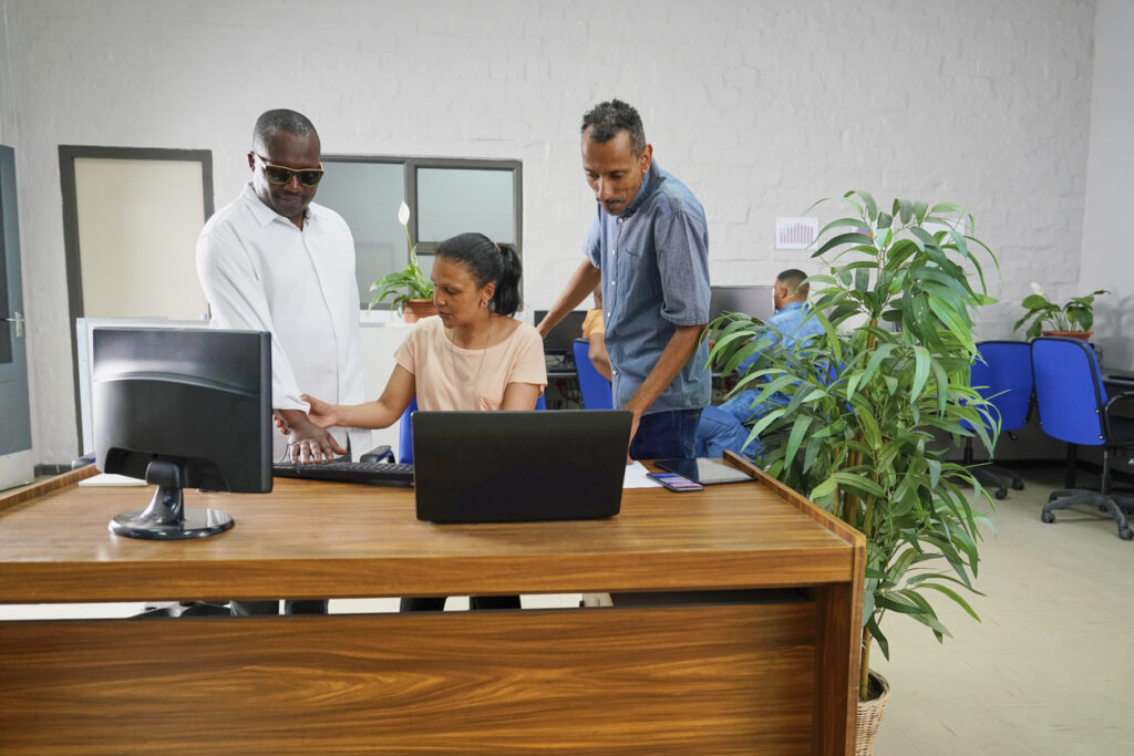 A person wearing dark glasses standing beside two coworkers as one guides their hand on a computer keyboard.