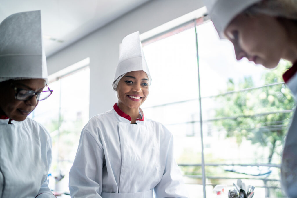 A person with a visual disability smiling while working in a chef’s uniform with coworkers in a commercial kitchen.