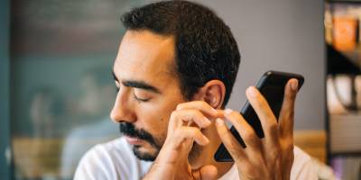 A person holding a smartphone close to their ear with eyes closed, listening to VoiceOver and using touch gestures while seated in their office.