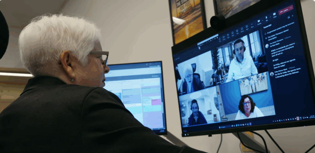 A person with partial sight sitting close to two large computer monitors at a desk while participating in a video meeting on one of the screens