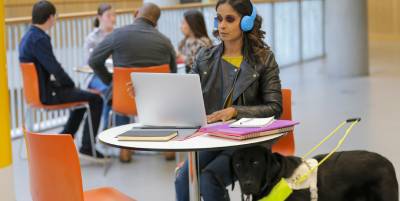 A person working on a laptop at a table with headphones on and a guide dog beside them, in a shared workspace with other people seated in the background