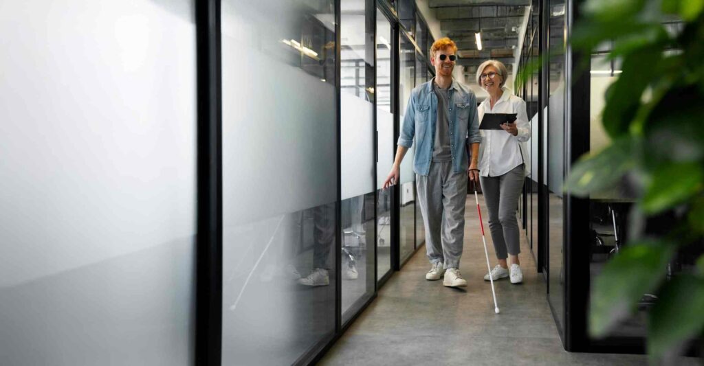 A person with a white cane and dark glasses walking through a bright office hallway beside a smiling coworker holding a tablet.