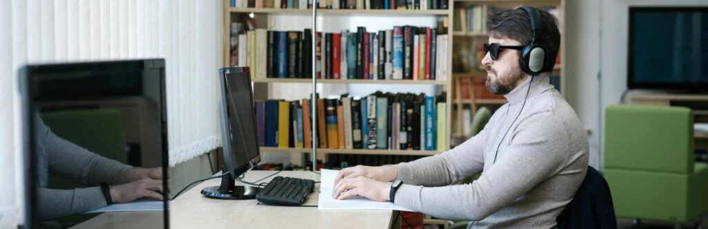 A person wearing dark glasses and headphones sitting at an office desk, reading braille with a computer and keyboard in front of them.